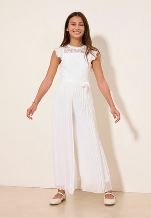 Joven chica sonriendo, vestida con una blusa sin mangas de encaje blanca con volantes y pantalones anchos plisados, de pie frente a una pared beige.