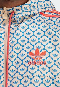 La chaqueta cortavientos de Adidas presenta un patrón hexagonal en azul y blanco con pequeños logotipos, un zipper rojo y un logotipo bordado en rojo en el pecho.