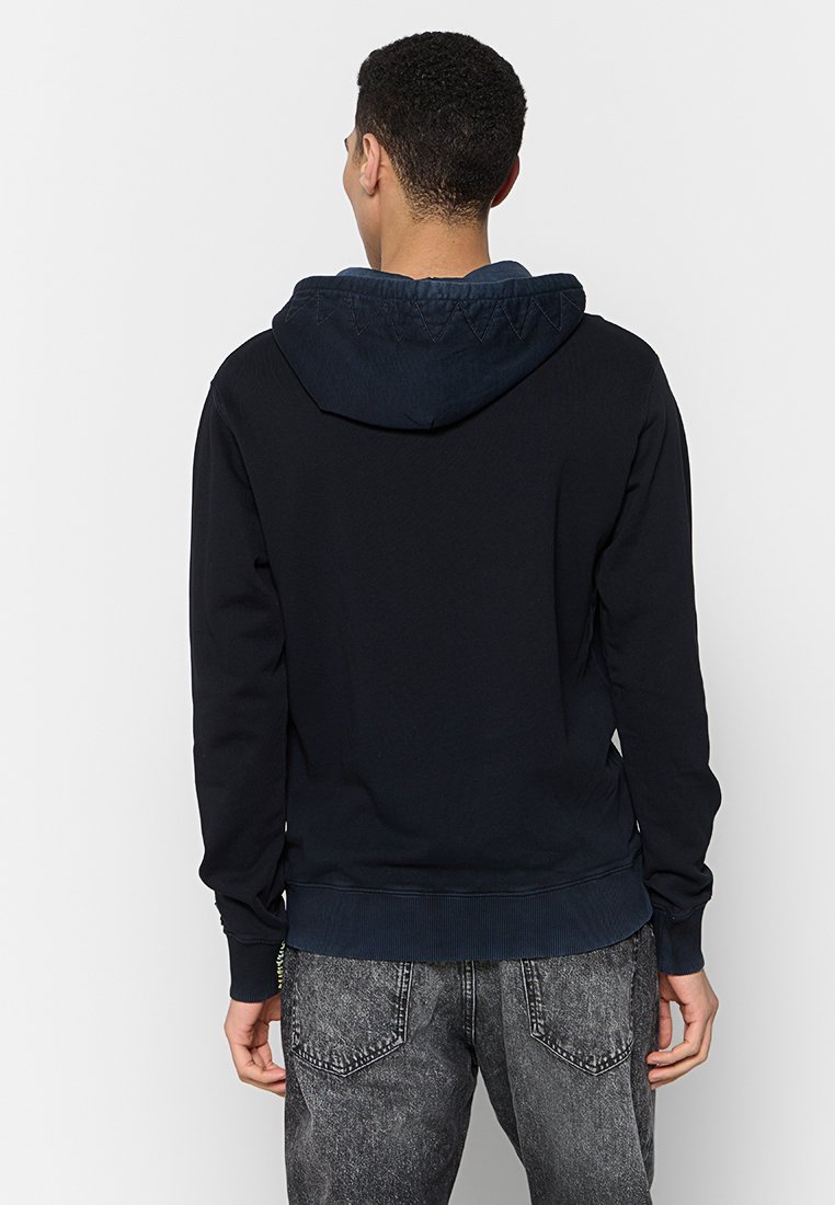 Ein Mann trägt einen dunkelblauen Hoodie mit herabgeklapptem Kapuzen und steht mit dem Rücken zur Kamera, kombiniert mit ausgebleichten schwarzen Jeans.