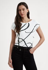 Baltas trumpomis rankovėmis marškinėlių viršus su abstrakčiu juodos linijos dizainu. Apvalus kaklo iškirpimas ir lygi faktūra. Dėvimas su juodos spalvos aukšto liemens džinsais ir diržu.