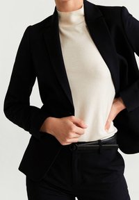 Černý blazer přes krémový rolák, doplněný černými kalhotami a úzkým páskem s kulatou sponou, který zdůrazňuje strukturovanou siluetu.