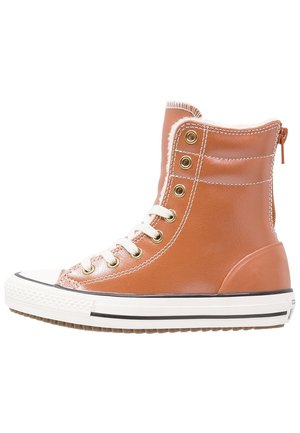 Brązowy wysokowy but sportowy z białymi sznurowadłami, złotymi przelotkami, białą gumową podeszwą z czarnymi paskami oraz widoczną kożuchową wyściółką przy kostce.