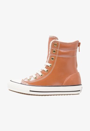 Zapatilla deportiva marrón de caña alta con cordones blancos, ojales dorados, suela de goma blanca con rayas negras y forro de borrego visible en el tobillo.