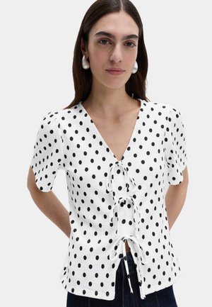 Hvid bluse med korte ærmer og sorte polkaprikker, der har en dyb V-hals og bindedetalje foran. Stoffet har en struktureret overflade.