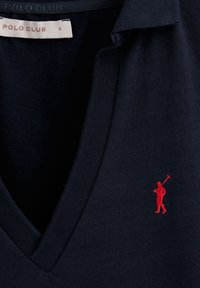 Marinblå pikétröja med V-halsdesign. Har en liten röd broderad logotyp av en poloåkare på vänster sida. Mjuk, strukturerad tyg.