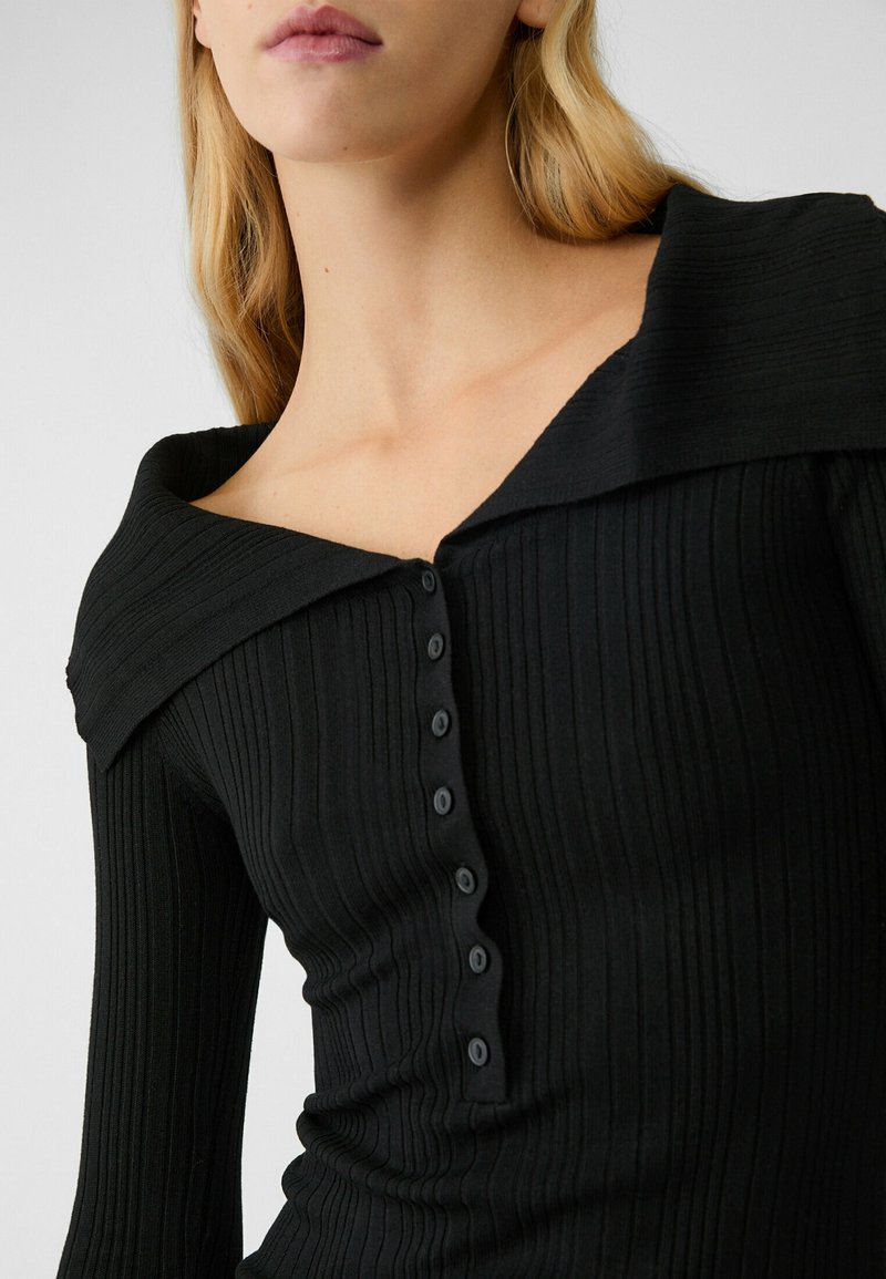Maglietta nera a maniche lunghe con costine, dal collo largo e scollato, con una fila di sei bottoni neri sul davanti. Tessuto dalla texture liscia.