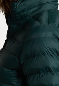 Veste matelassée verte avec un col montant, un design matelassé et une texture lisse. Comprend une fermeture éclair partielle et un petit détail de logo.