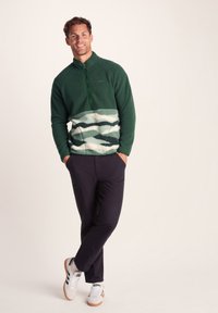 Green fleece pullover with a zip collar, featuring a light green and white wave pattern at the waist. Worn with dark pants and white sneakers.