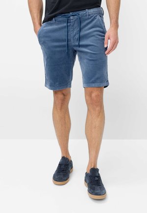 Mann trägt blaue Cord-Shorts mit Kordelzug in der Taille, marineblaue Sneakers und ein schwarzes T-Shirt, steht vor weißem Hintergrund.
