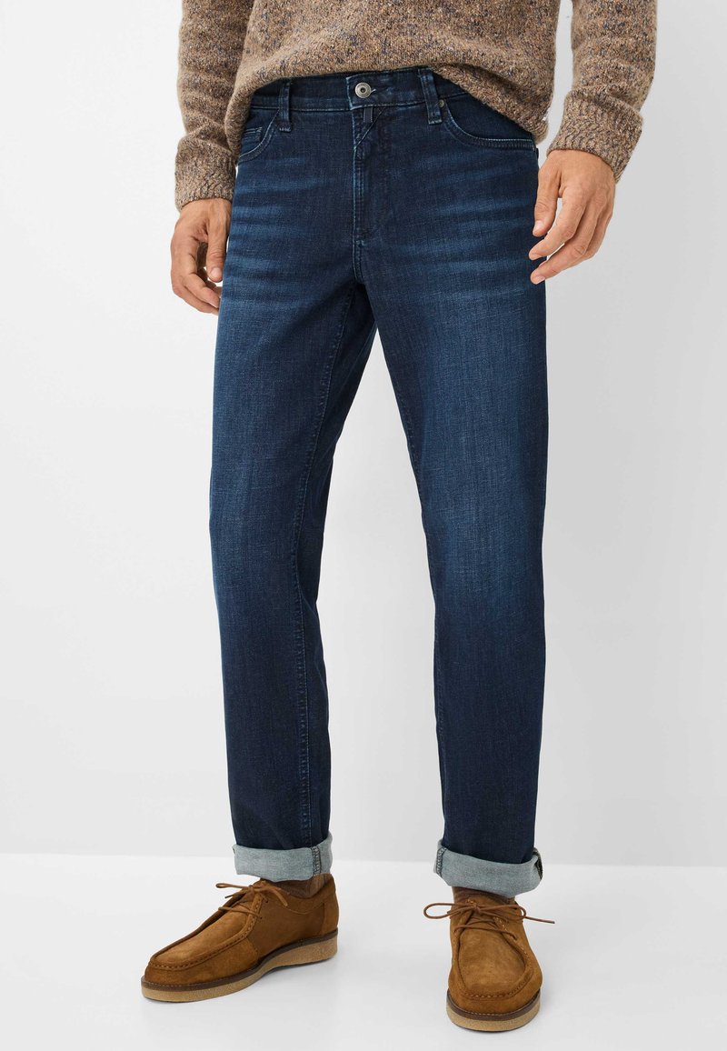 Jeans in denim blu scuro con taglio dritto, dotati di apertura con bottoni, cinque tasche e leggeri sbiadimenti sulle cosce. I risvolti mostrano scarpe beige.