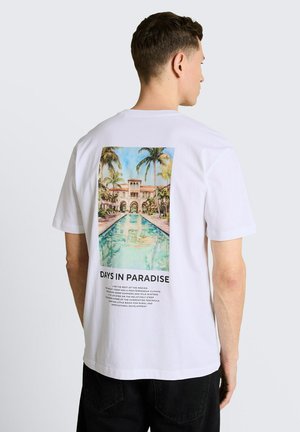 Man wearing white T-shirt with a colorful print of a palm-lined pool and Mediterranean-style building, text reads "DAYS IN PARADISE."