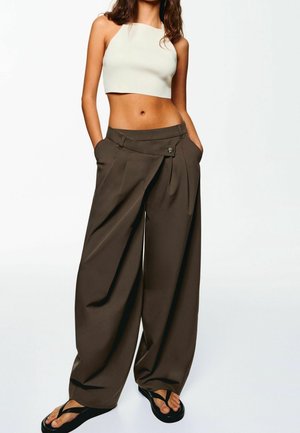 Kvinde iført en hvid cropped tank top, løse brune vidde bukser med folder og sorte flip-flops, står med hænderne i lommerne.
