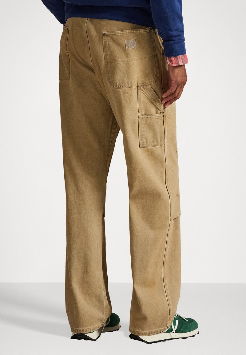 Pantaloni da lavoro beige realizzati in tessuto resistente, con molteplici tasche, una vestibilità ampia e una cintura rinforzata. Visti da dietro.