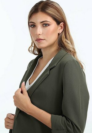 Grüne Blazer mit tailliertem Design, ausgestattet mit einem Reverskragen und langen Ärmeln. Glattes Material und eine weiße Innenschicht, die am Ausschnitt sichtbar ist.