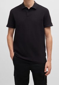 Polo noir en coton, avec un col court, une patte de boutonnage à deux boutons et un logo discret. Texture lisse, design ajusté et manches roulées.