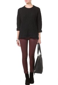Mujer vistiendo un suéter negro texturizado, pantalones de cuero burdeos, botas negras hasta el tobillo, sosteniendo un gran bolso negro por las asas.