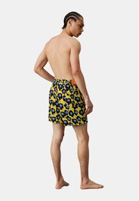 Pantaloni da bagno con una base gialla e un motivo floreale blu, progettati con una tasca sul retro e un'etichetta arancione. Tessuto morbido con colori vivaci.