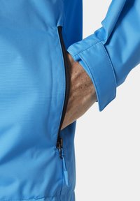 Chaqueta azul claro con un bolsillo con cremallera. Presenta una textura suave y ligeramente brillante, y tableros ajustables en los puños. Adornada con acentos de cremallera negra.