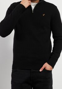 Pull noir zippé en maille texturée, avec un ourlet et des poignets côtelés, et un petit logo brodé sur la poitrine.