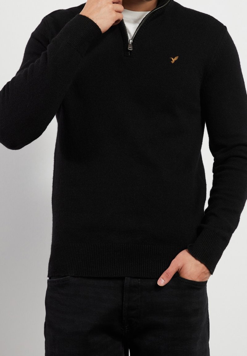 Pull noir zippé en maille texturée, avec un ourlet et des poignets côtelés, et un petit logo brodé sur la poitrine.