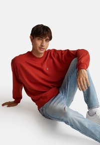 Felpa rossa di cotone con polsini e scollatura a costine, con un piccolo logo ricamato blu, abbinata a jeans in denim blu chiaro e sneakers bianche.