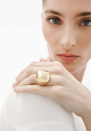 Anillo de color dorado con una parte superior cuadrada lisa y redondeada, llevado en un dedo. La mano del modelo reposa sobre el hombro, con un fondo blanco liso.