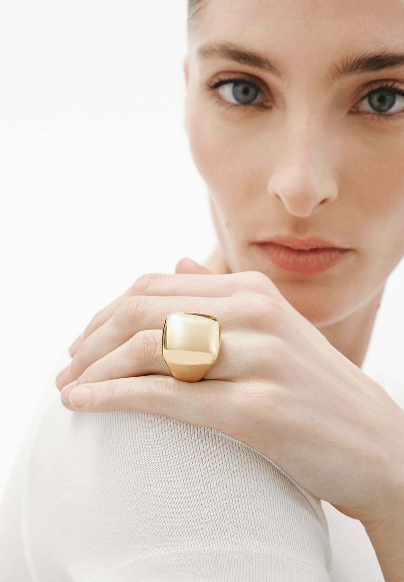 Anillo de color dorado con una parte superior cuadrada lisa y redondeada, llevado en un dedo. La mano del modelo reposa sobre el hombro, con un fondo blanco liso.