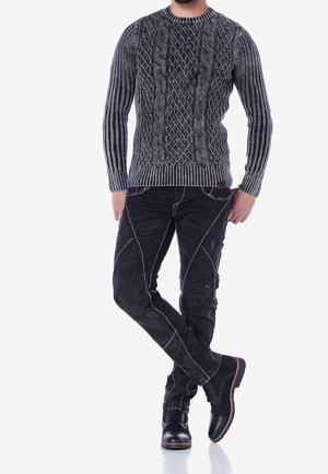 Mann trägt einen strukturierten grauen Zopfpullover, schwarze ausgewaschene Jeans mit weißen Nähten und schwarze Lederstiefel, steht mit überkreuzten Beinen.