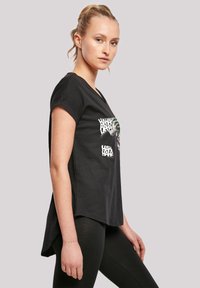 Svart t-skjorte med korte ermer, med et grafisk trykk i hvitt og grønt. Designet har en avslappet passform og en lengre bakdel.