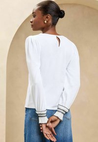 Witte blouse met lange mouwen en een sleutelgatdetail op de achterkant, geraffineerde mouwen en zwarte trim op de elastische manchetten. Gecombineerd met blauwe spijkerbroek.
