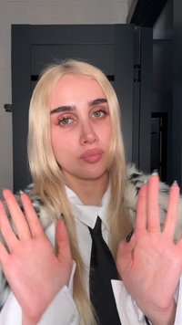 Jong persoon met lang blond haar, gekleed in een wit shirt, zwarte stropdas en een patroonjas, die beide handen naar voren opheft binnenshuis.