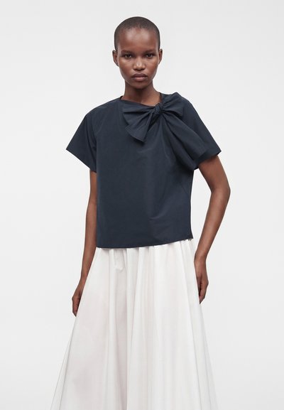 Femme portant un haut noir à manches courtes avec un grand nœud à l'épaule et une jupe blanche fluide sur un fond uni.