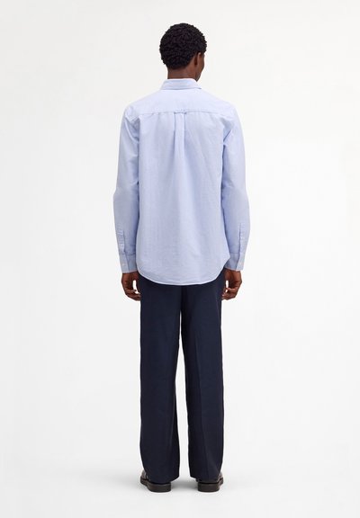 Mężczyzna stojący tyłem, ubrany w jasnoniebieską, paskowaną koszulę z długim rękawem, ciemnogranatowe spodnie i czarne buty na białym tle.