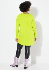 Langärmliges, leuchtend limonengrünes Tunika mit abgerundetem Saum, kombiniert mit schwarzen Leggings und glänzenden, silbernen Cowboystiefeln mit spitzer Zehenpartie.