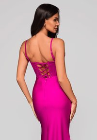 Femme aux longs cheveux noirs portant une robe rose vif moulante avec un détail laçé dans le dos, debout devant un fond gris clair.