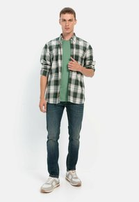 Grünes T-Shirt, das unter einem schwarz-weißen karierten Hemd mit Knöpfen getragen wird, kombiniert mit blauen Jeans und beigen Sneakers mit grauen Akzenten.