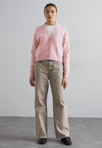 Lichtroze cardigan met een V-hals, geribbelde rand en knopen, gecombineerd met beige wijde jeans en zwarte schoenen. Eenvoudige, casual outfit.