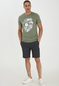 Camiseta de algodón verde con gráfico de palmera blanca y texto, combinada con pantalones cortos oscuros y zapatillas blancas. Fondo plano.