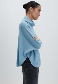 Frau mit dunklem Haar im Dutt, die silberne Ohrringe, einen lockeren hellblauen Rollkragenpullover und dunkle Jeans trägt, mit verschränkten Armen.