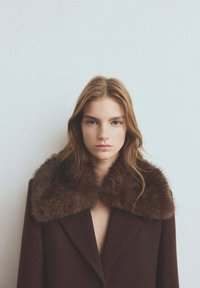 Bruine wollen jas met een grote, zachte bontkraag en een diep V-hals ontwerp. De stof lijkt glad te zijn met een gestructureerde silhouet.