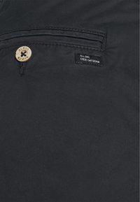 Tissu en denim noir présentant une surface texturée, un bouton en bois et une petite étiquette avec "DENIM TOUS LES JOURS, TOUTE LA JOURNÉE" brodée en blanc.