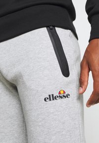 Grey cotton joggers feature a black zipper pocket and an "ellesse" logo with a red, yellow, and black accent on the thigh.