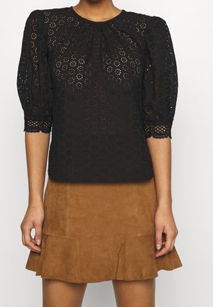 Vrouw die een zwarte blouse met kantpatroon en halflange mouwen draagt, gecombineerd met een bruine suède minirok met wijde zoom.