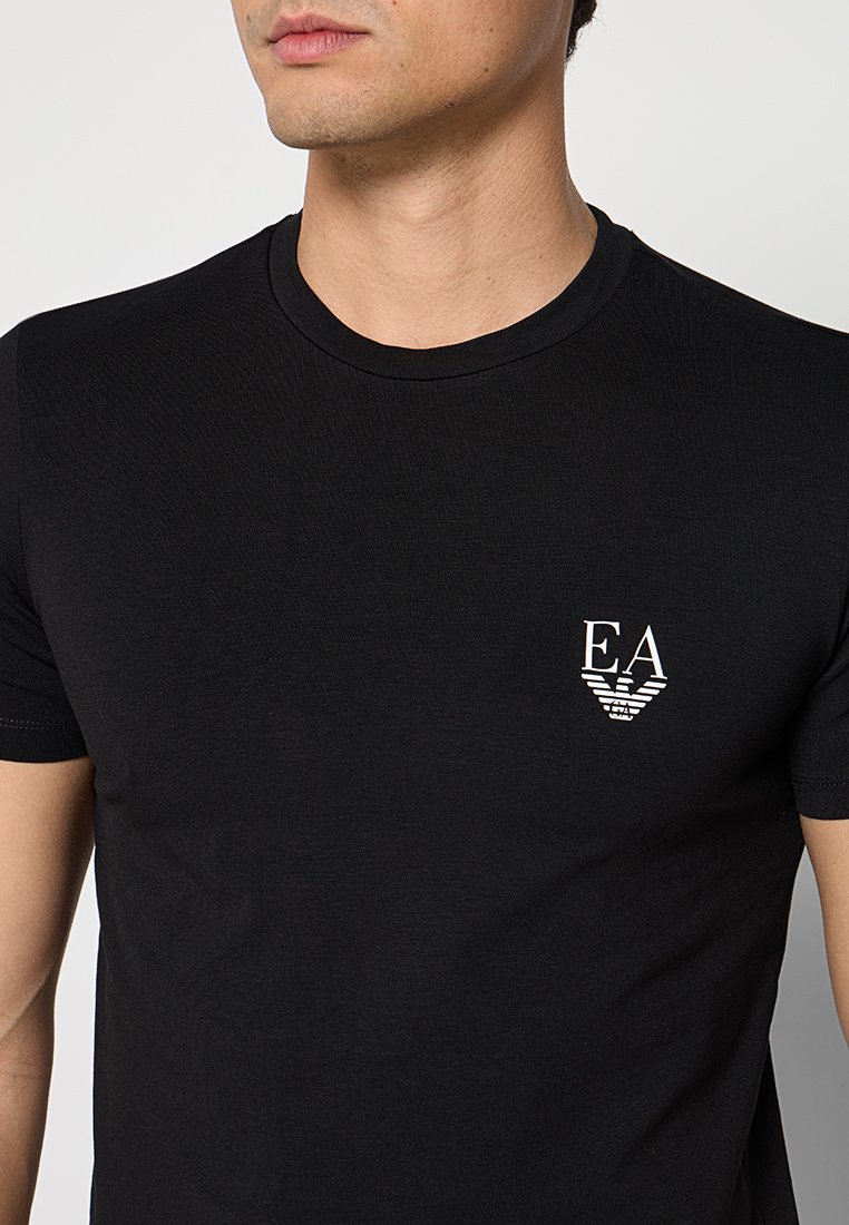 Hombre con una camiseta negra de cuello redondo con el logo blanco "EA" y un águila en el pecho izquierdo.