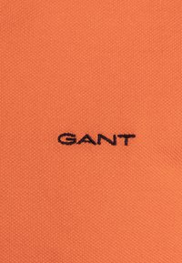 Oranžová pletená tkanina s texturovaným povrchem, v jejímž středu je černě vyšívané logo "GANT".