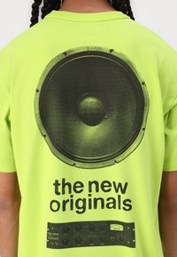 Neon grønn t-skjorte med et svart høyttalerbilde og tekst "de nye originalene" over et svart grafisk equalizer-design.
