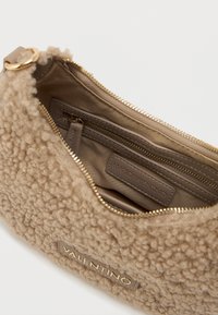 Bežs tekstaturs soma ar mīkstu, pūkainu ārpusi, zelta rāvējslēdzis un iekšējo nodalījumu ar etiķeti un papildu kabatu.