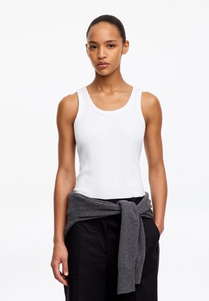 Jeune femme aux cheveux attachés en arrière, portant un haut blanc sans manches et un pantalon noir avec un pull gris noué autour de la taille.