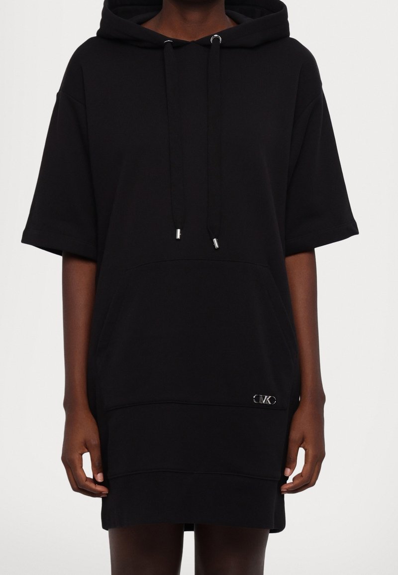 Personne portant une robe à capuche noire ample à manches courtes avec poche avant et petit logo métallique près de l'ourlet.