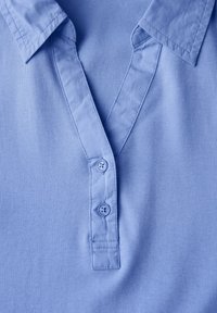 Svetle modré polo tričko s límečkovým výstřihem, dvěma knoflíky a jemnou, hladkou texturou. Materiál se zdá být lehký a neformální.
